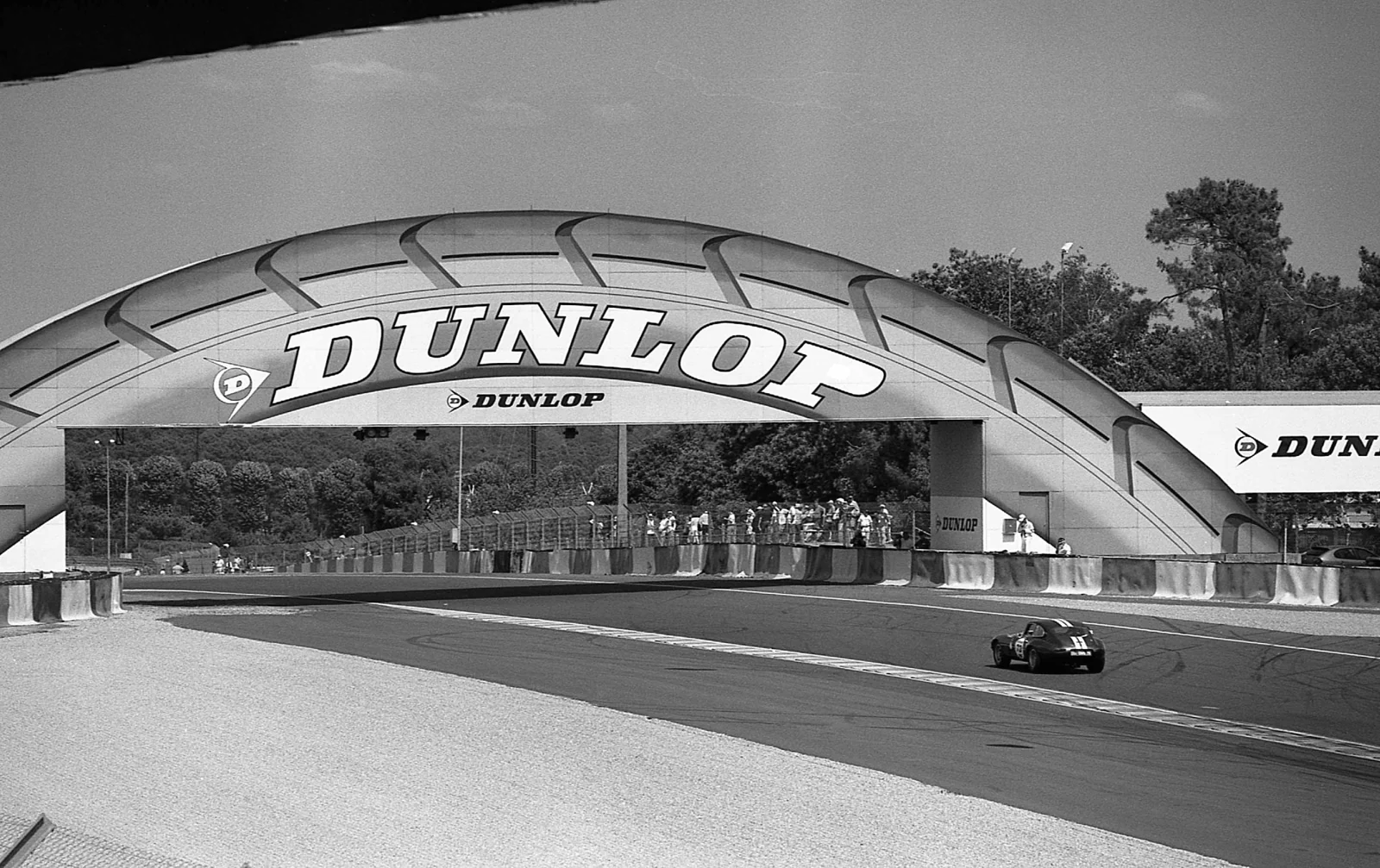 A black-and-white view of a race car beneath a circuit bridge.